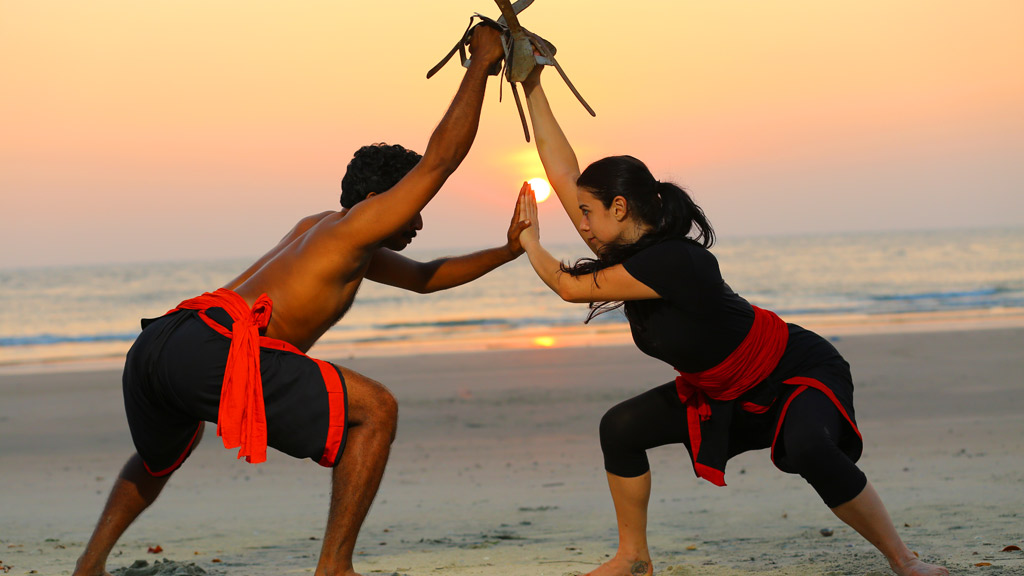 Two Kalaripayattu practitioners spar on a beach at sunset, wearing black training attire with red waist sashes. One holds traditional training weapons overhead while both maintain low martial stances, showcasing the agility, discipline, and techniques of Kerala's ancient martial art, Kalaripayattu.